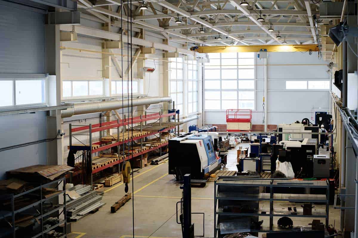 This photograph showcases a large, brightly lit factory floor, featuring an array of heavy machinery and organized storage racks. The scene is dominated by the gray concrete floor marked with yellow lines,